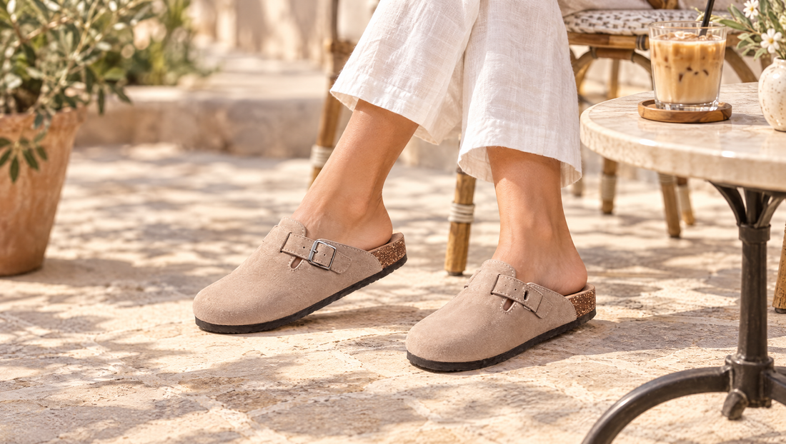 Voeten in taupe clogs van vegan velours met een witte linnen broek op een zonnig terras