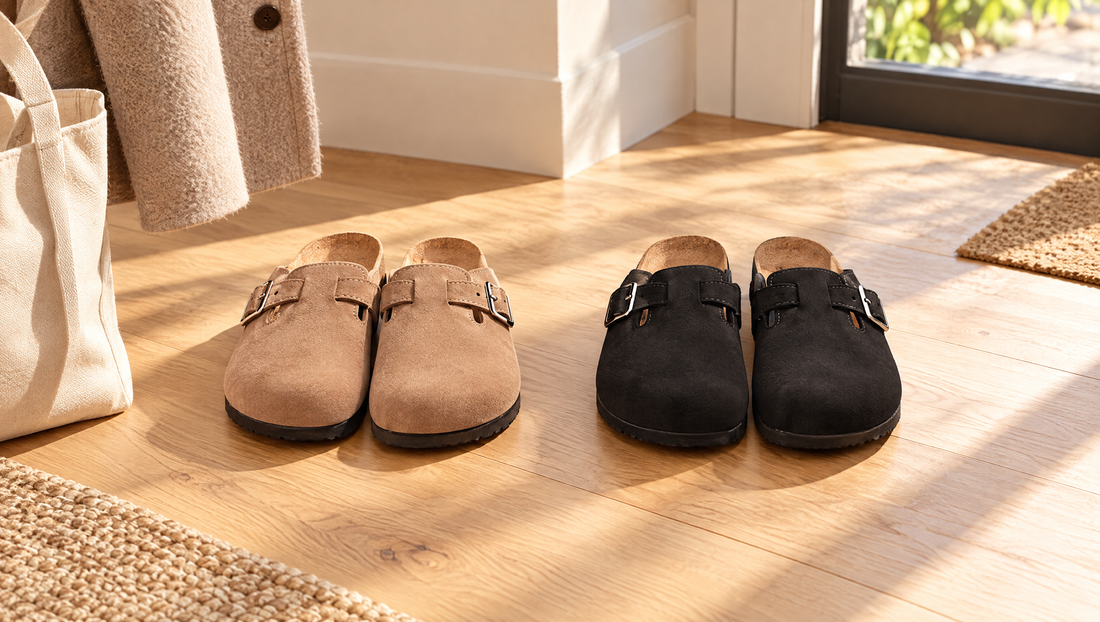 Twee paar clogs van vegan velours, camel en zwart, naast elkaar bij een deur op een zonnige houten vloer
